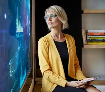 A businesswoman wearing a yellow blazer looking at analytical data screens in a modern corporate office.
